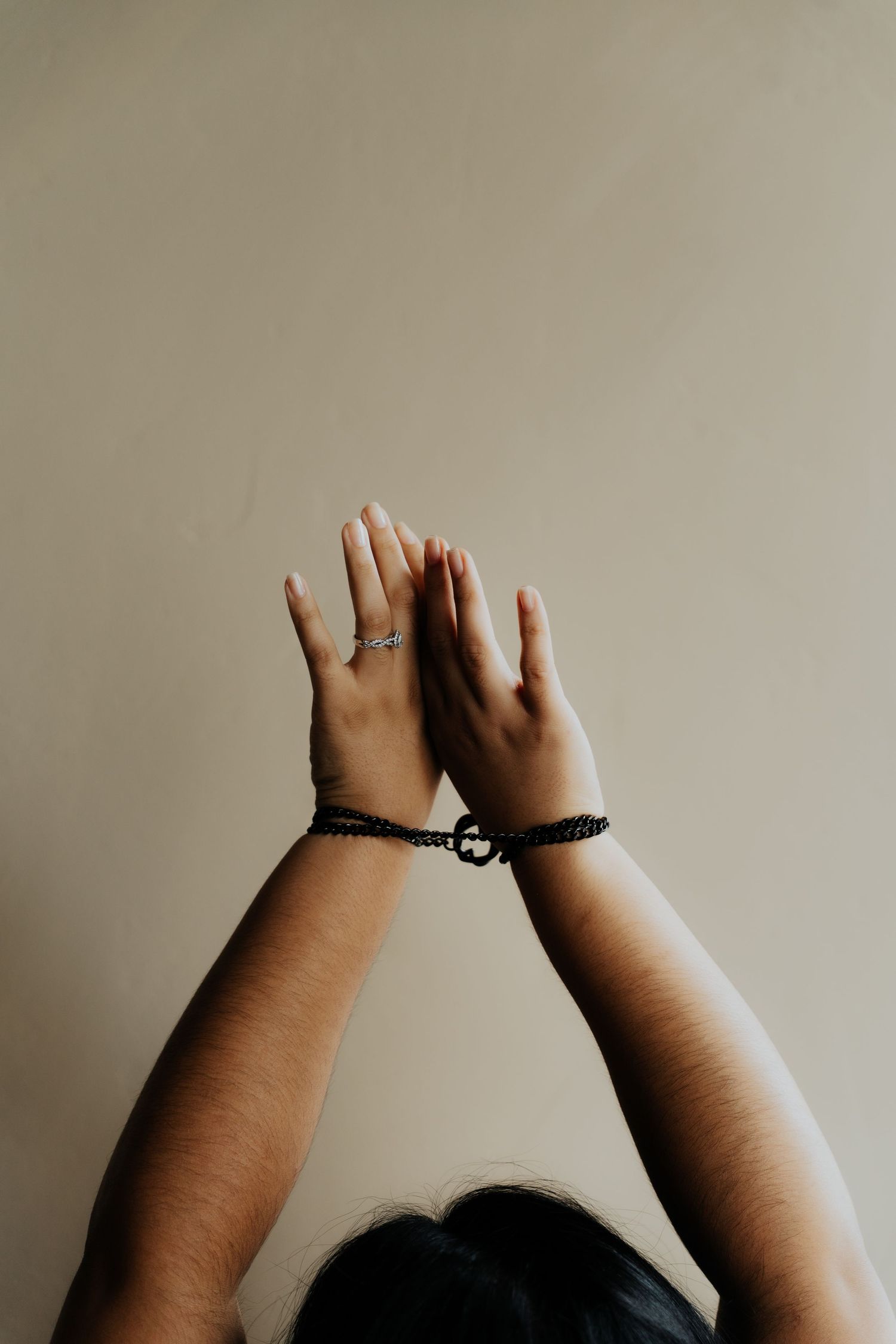 Two hands raised upward with wrists bound by a black bracelet against a beige wall.