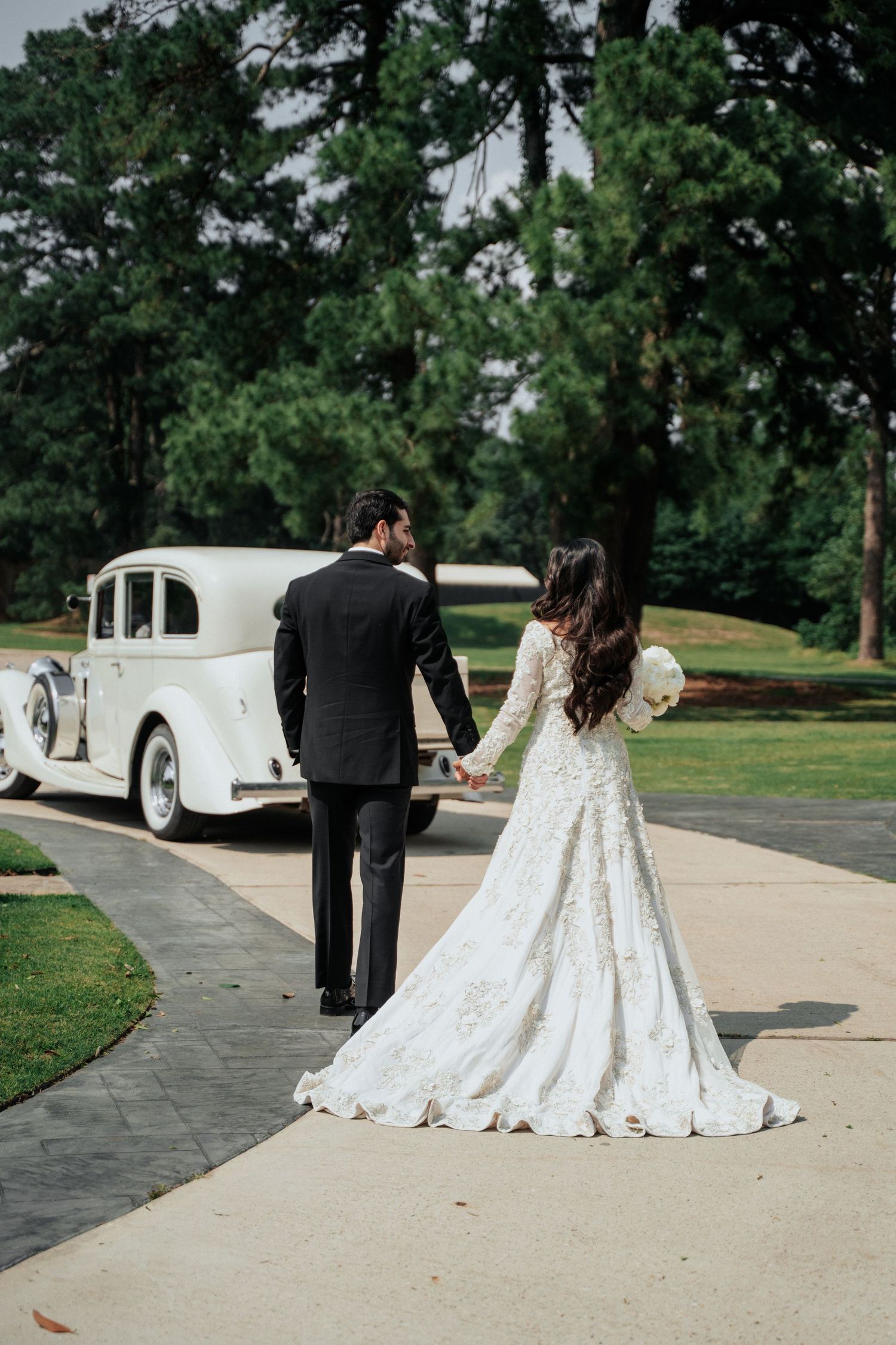 A wedding couple walks towards a classic white vintage car on a tree-lined path.