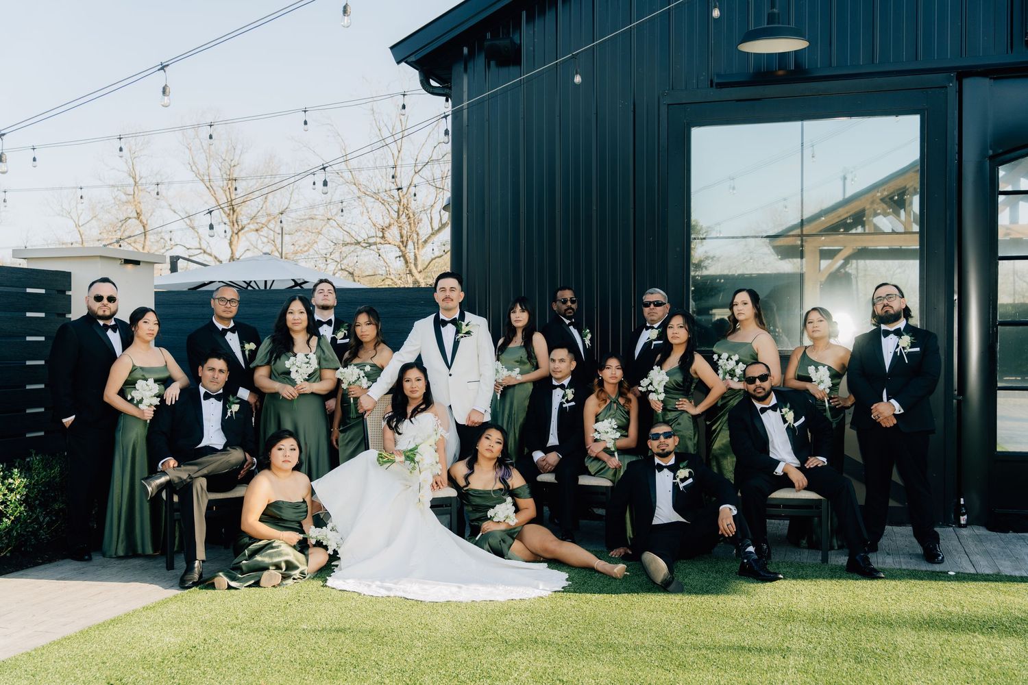 Wedding party poses together outdoors in formal attire with bridesmaids in dark dresses and groomsmen in tuxedos.