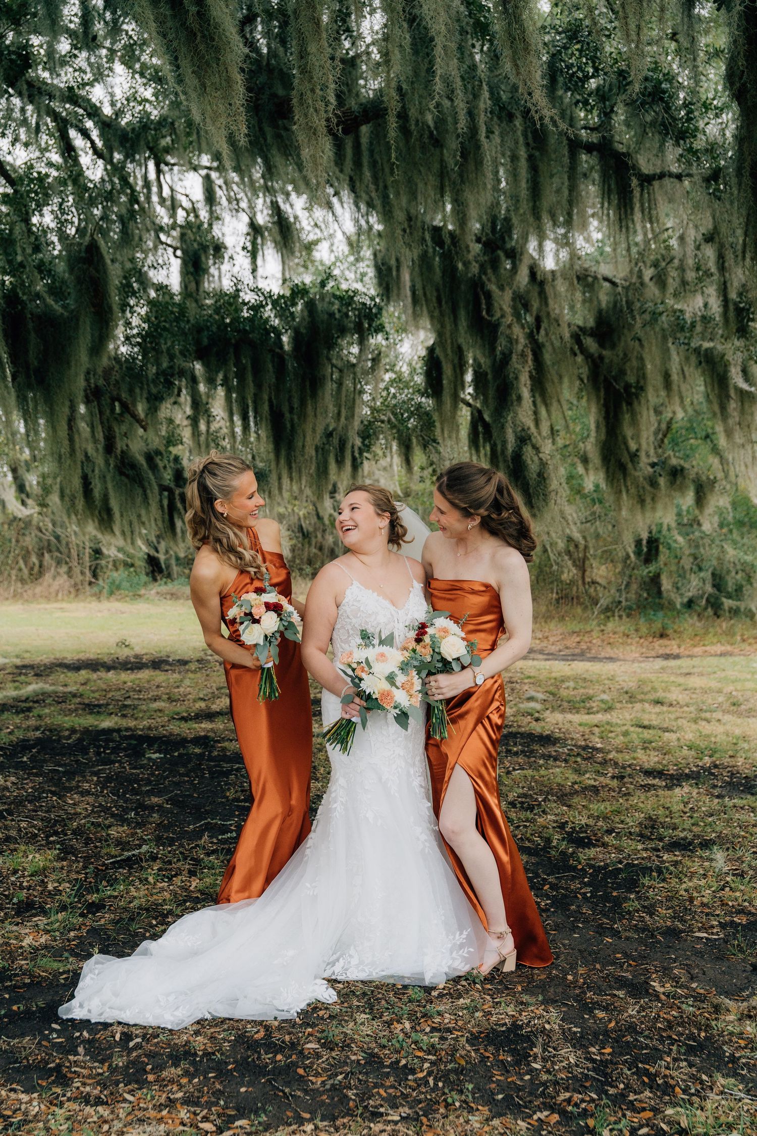 A wedding party poses under Spanish moss trees in a scenic outdoor setting.