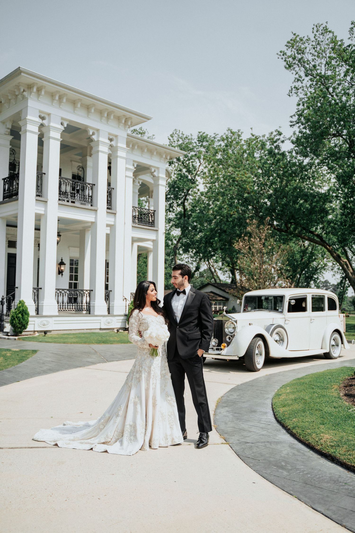 Elegant wedding couple poses outside grand white mansion with vintage car.
