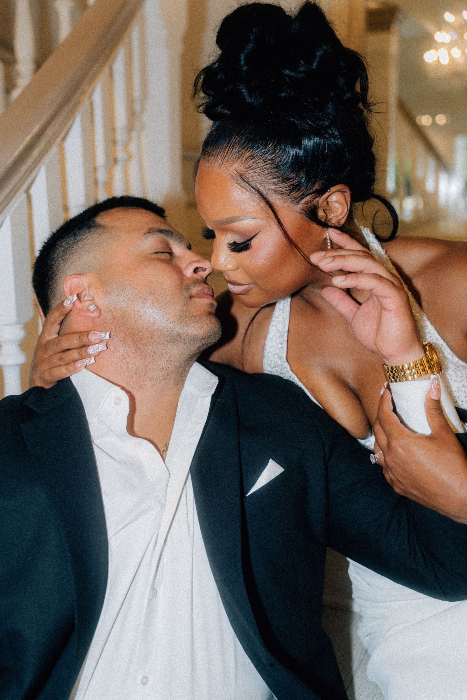 A couple shares a romantic almost-kiss moment, dressed in elegant wedding attire near a grand staircase.
