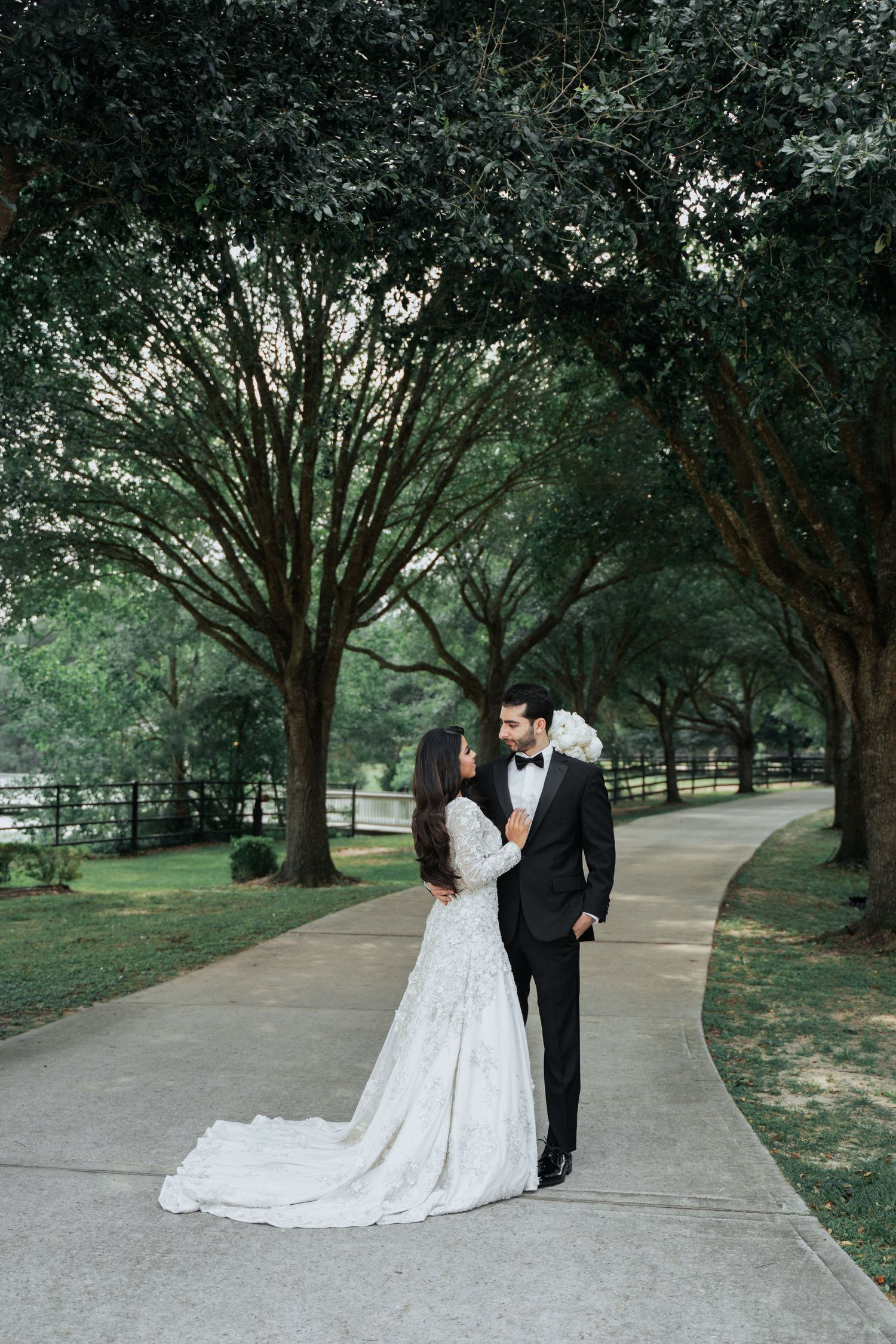 Wedding couple walking along tree-lined pathway in formal attire.