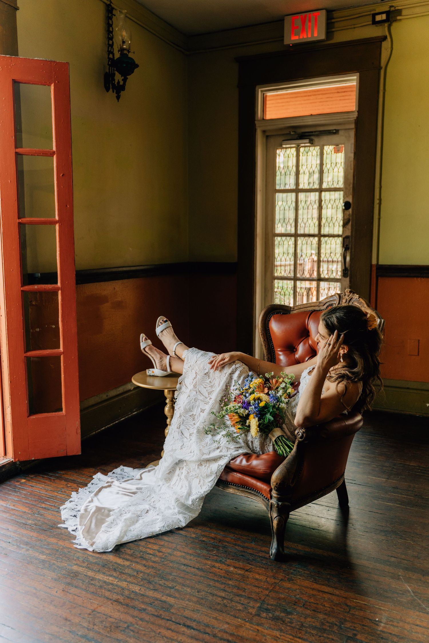 A bride in a lace wedding dress lounges on a vintage couch in a rustic room with wooden floors.