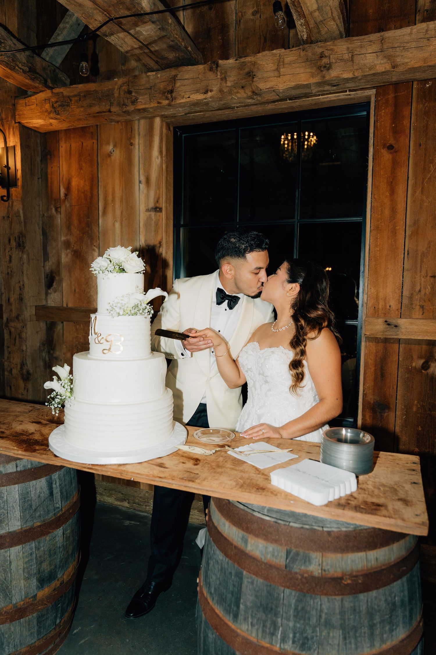 Elegant white wedding cake displayed on rustic wooden wine barrels in a barn setting.