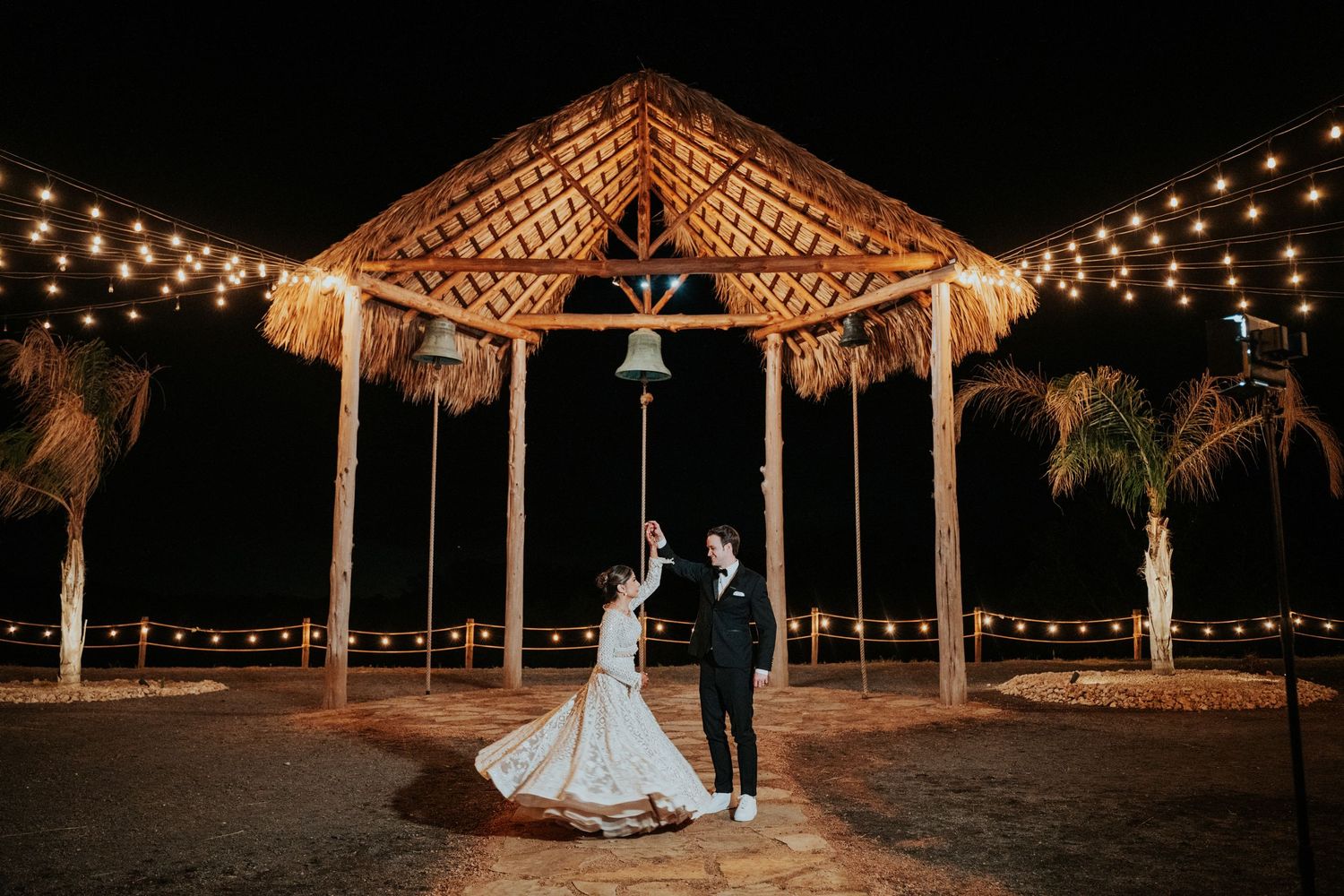 Romantic outdoor wedding venue at night with string lights illuminating palm trees and a gazebo.