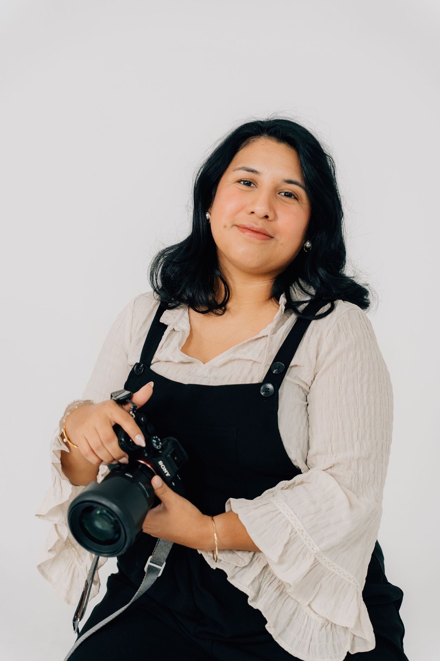 Photographer in white blouse and black overalls poses with camera equipment.