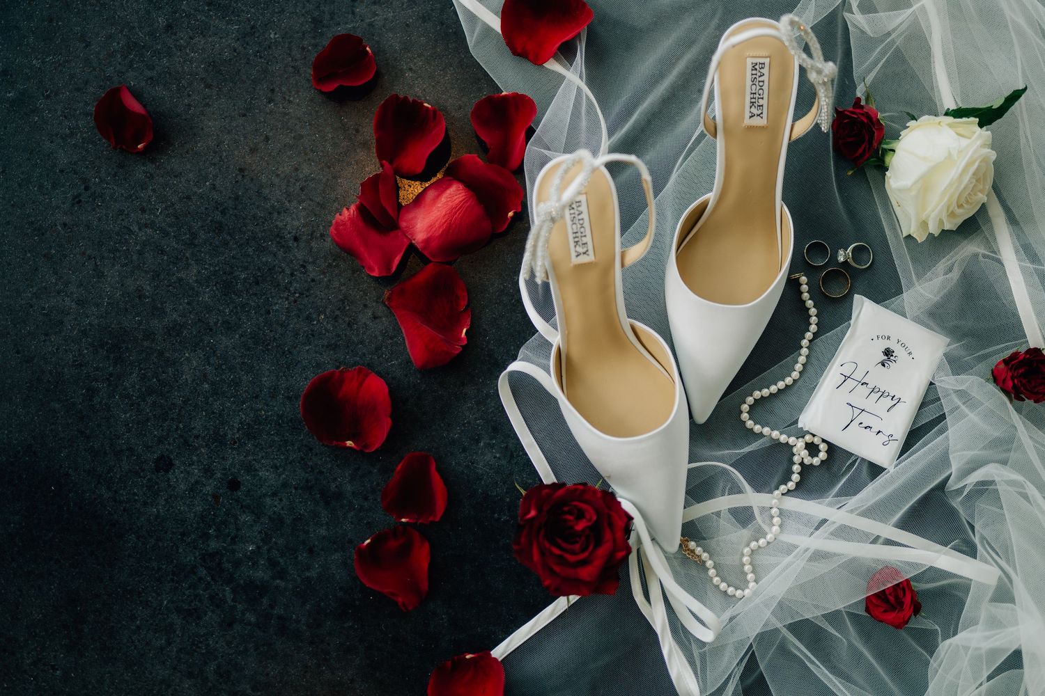 Wedding accessories including white shoes and scattered red rose petals against dark background.