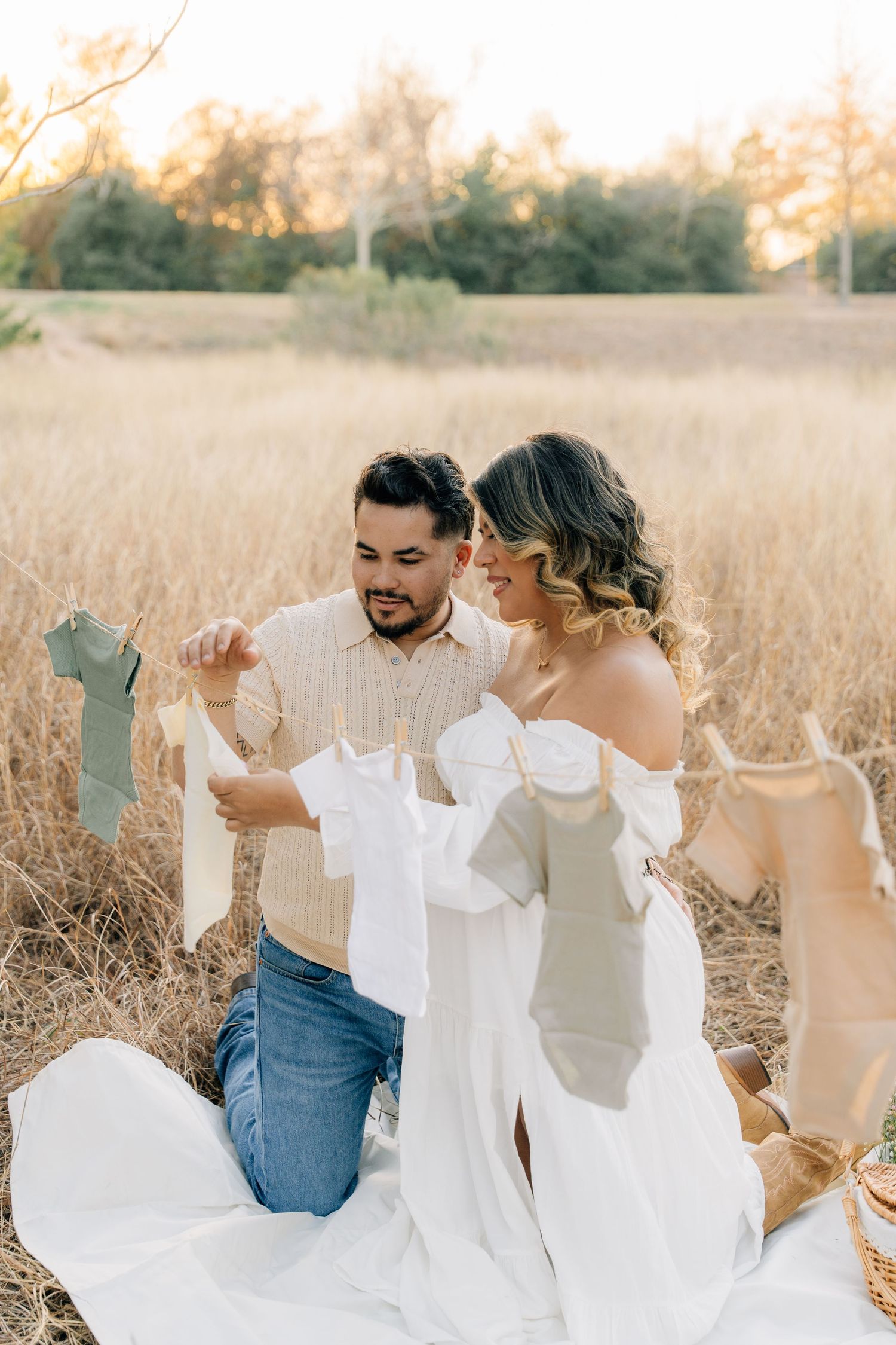 Couple in white outfits hanging baby clothes on clothesline in golden field at sunset.