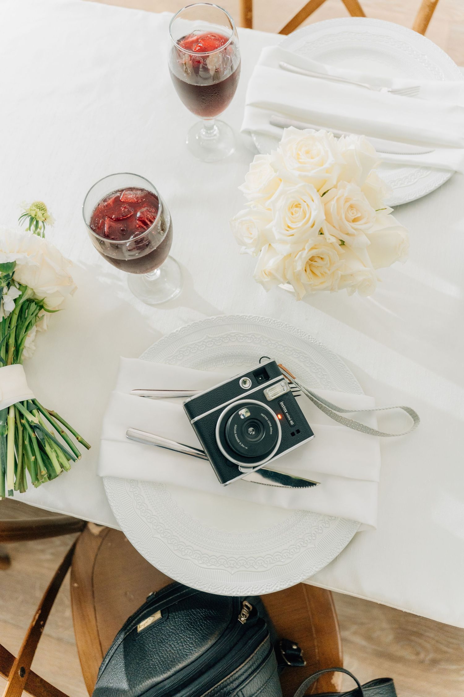 Table setting with wine glasses, white roses, and vintage camera on white tablecloth.