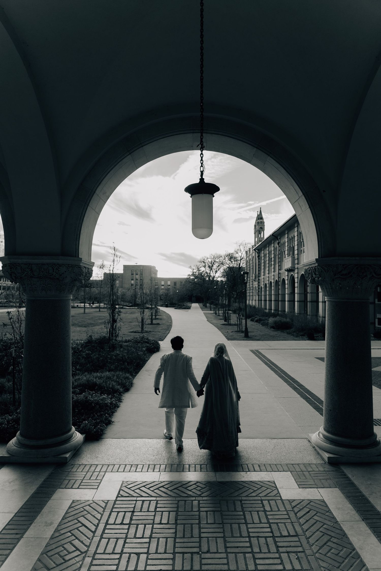 Black and white architectural photo of people walking through arched colonnade with geometric floor pattern.