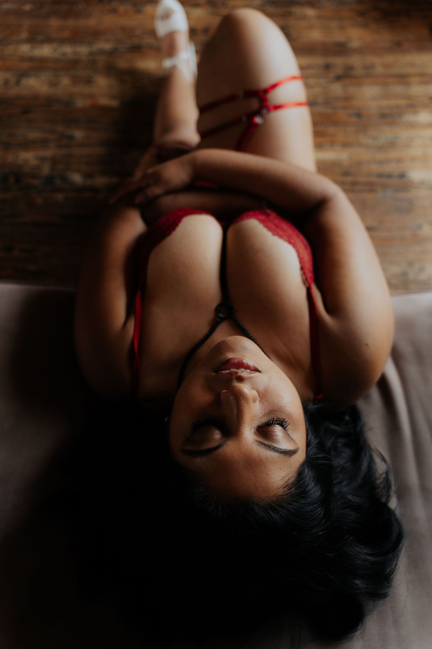 Artistic boudoir portrait of person lying on wooden surface with red lingerie details in moody lighting.