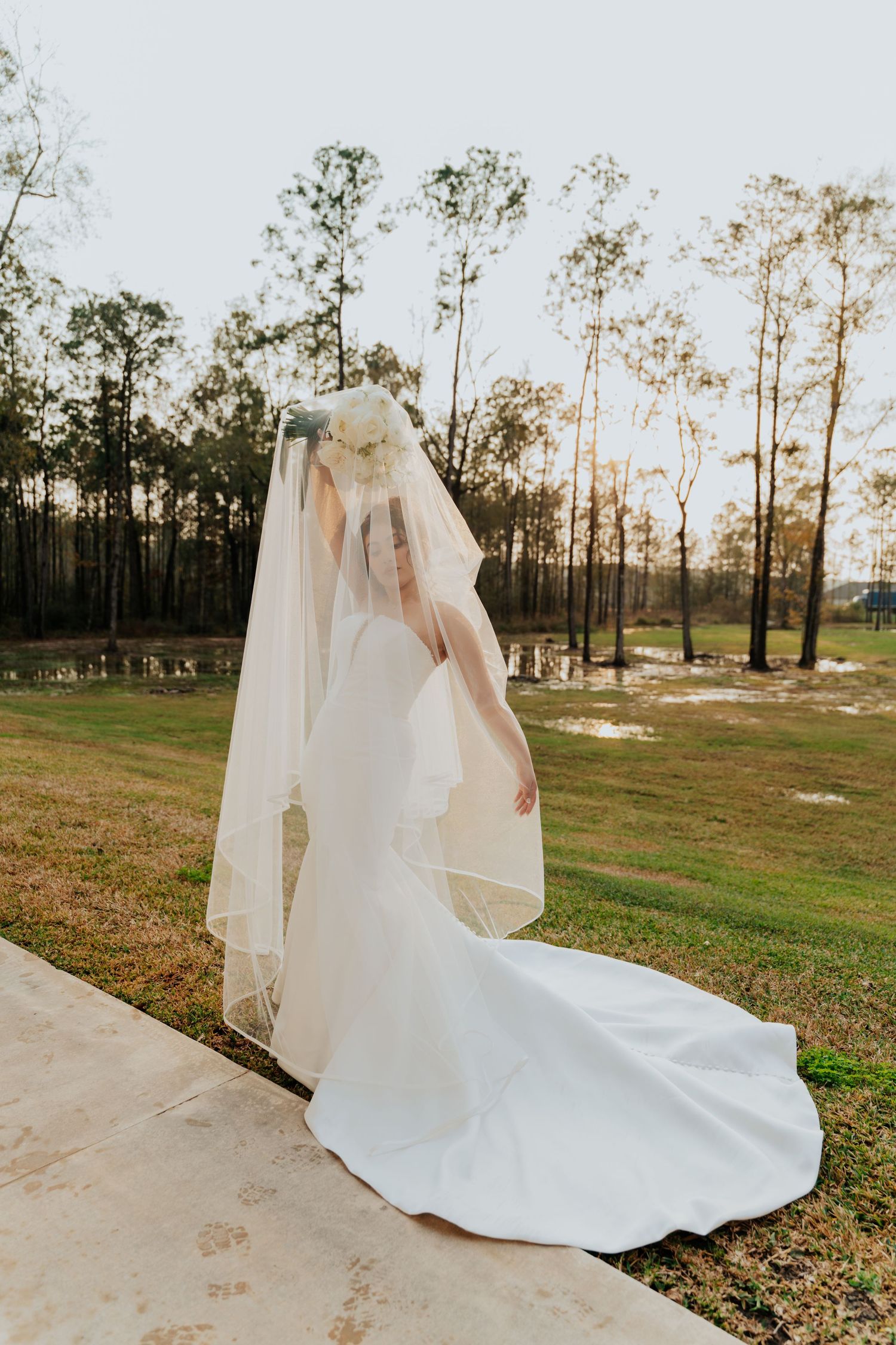 A flowing white wedding dress and veil captured in motion across a grassy field at sunset with tall trees in the background.