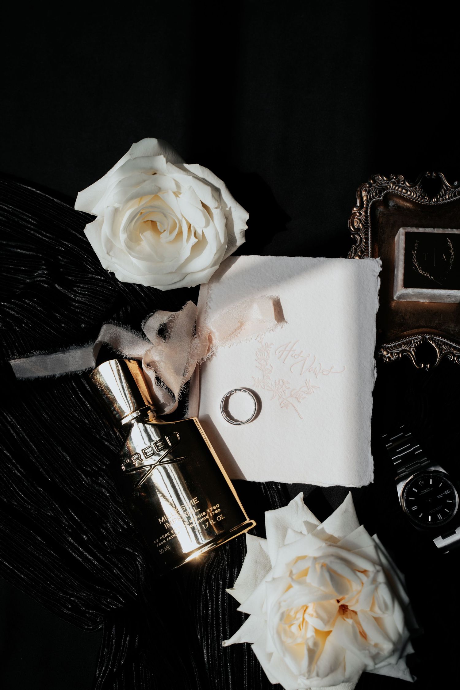 Elegant wedding details featuring white roses, rings, and perfume bottle on dark marble surface.