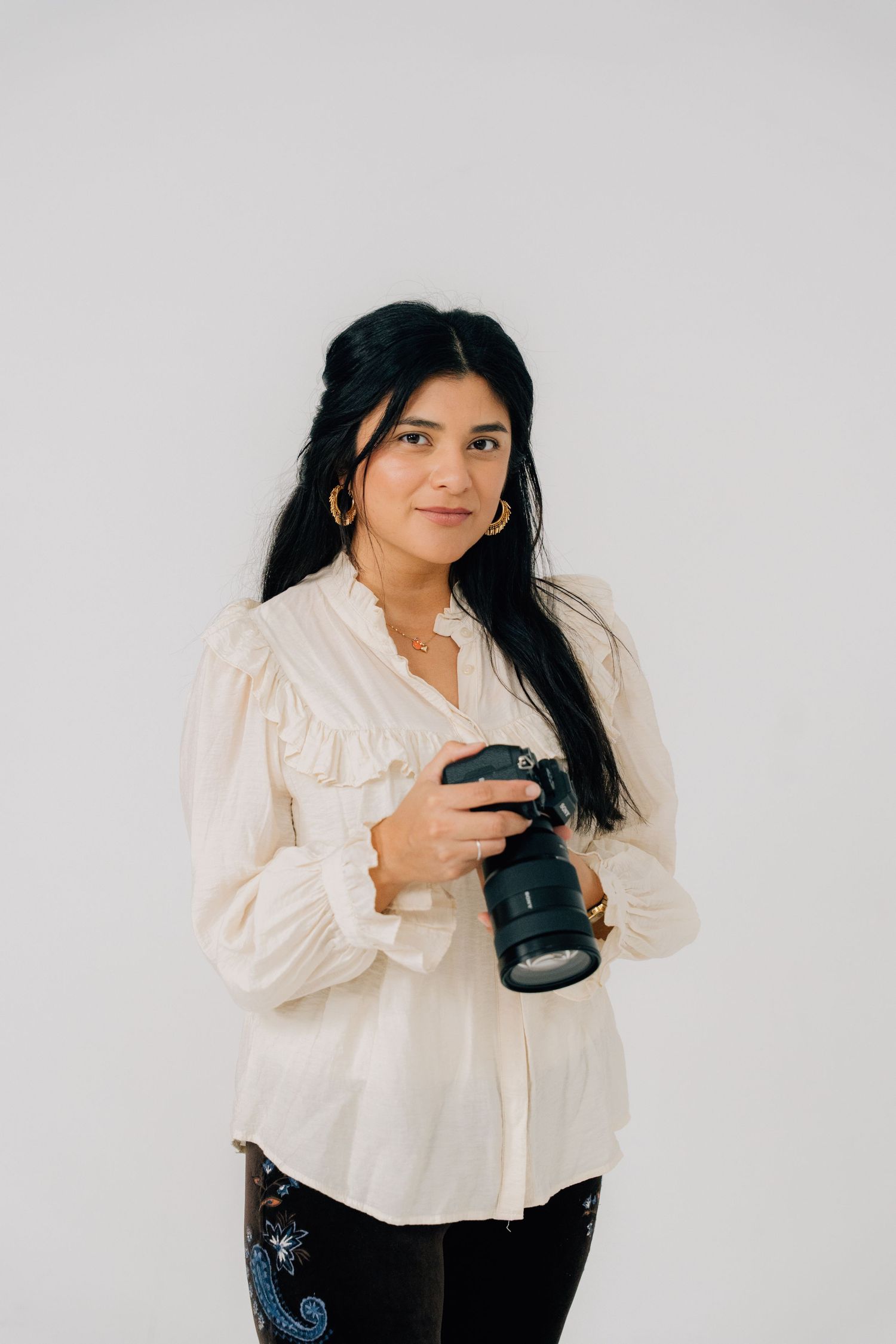 Photographer in white ruffled blouse holds camera against white studio background.