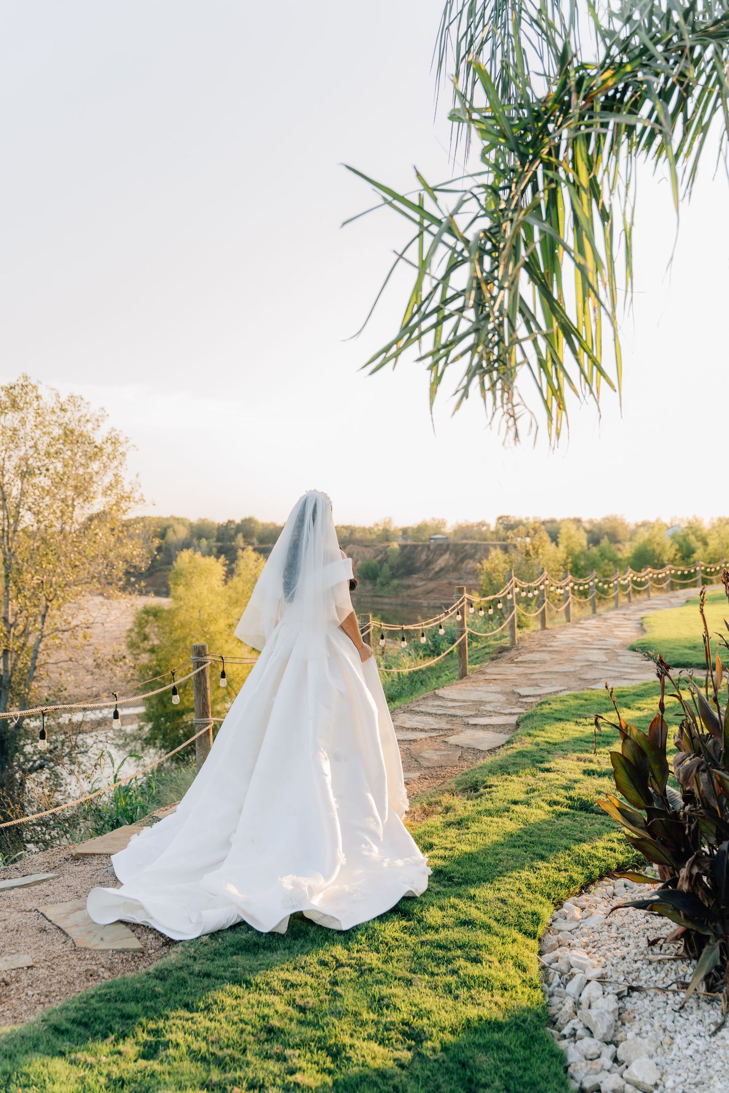 A bride in a flowing white wedding dress and veil walks along a scenic garden path on a sunny autumn day.