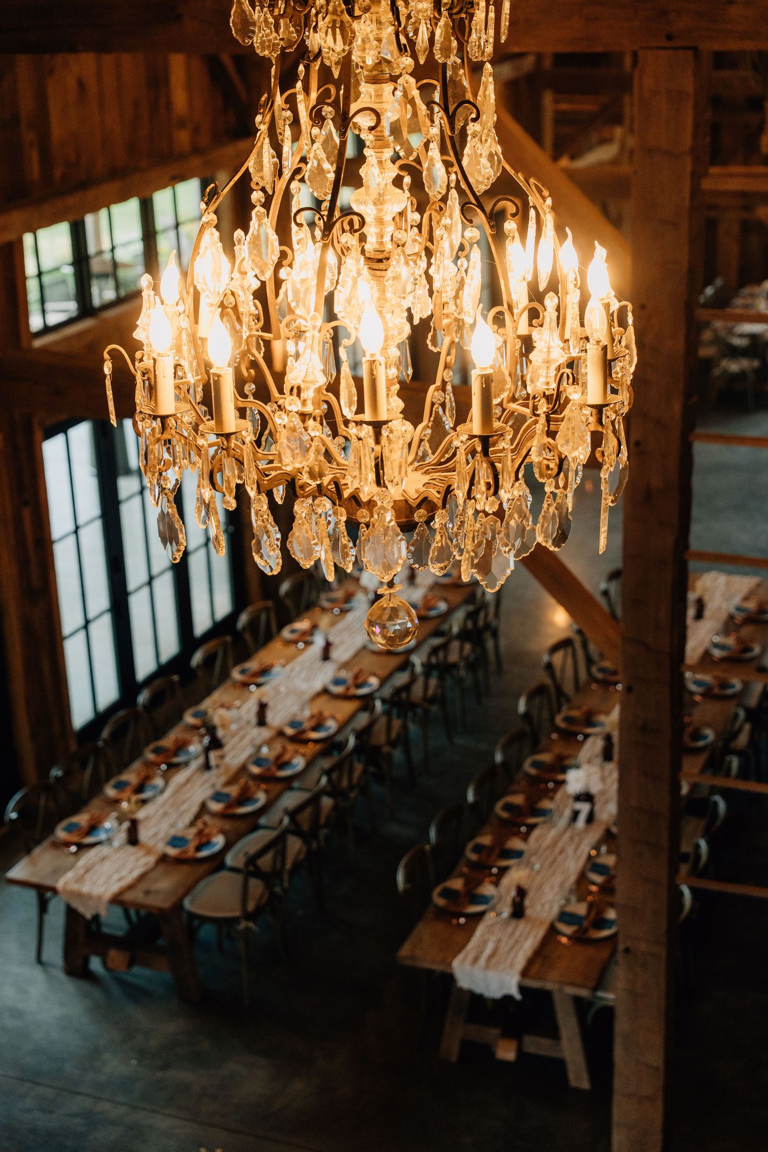 Elegant crystal chandeliers hanging over long reception tables in a rustic venue.
