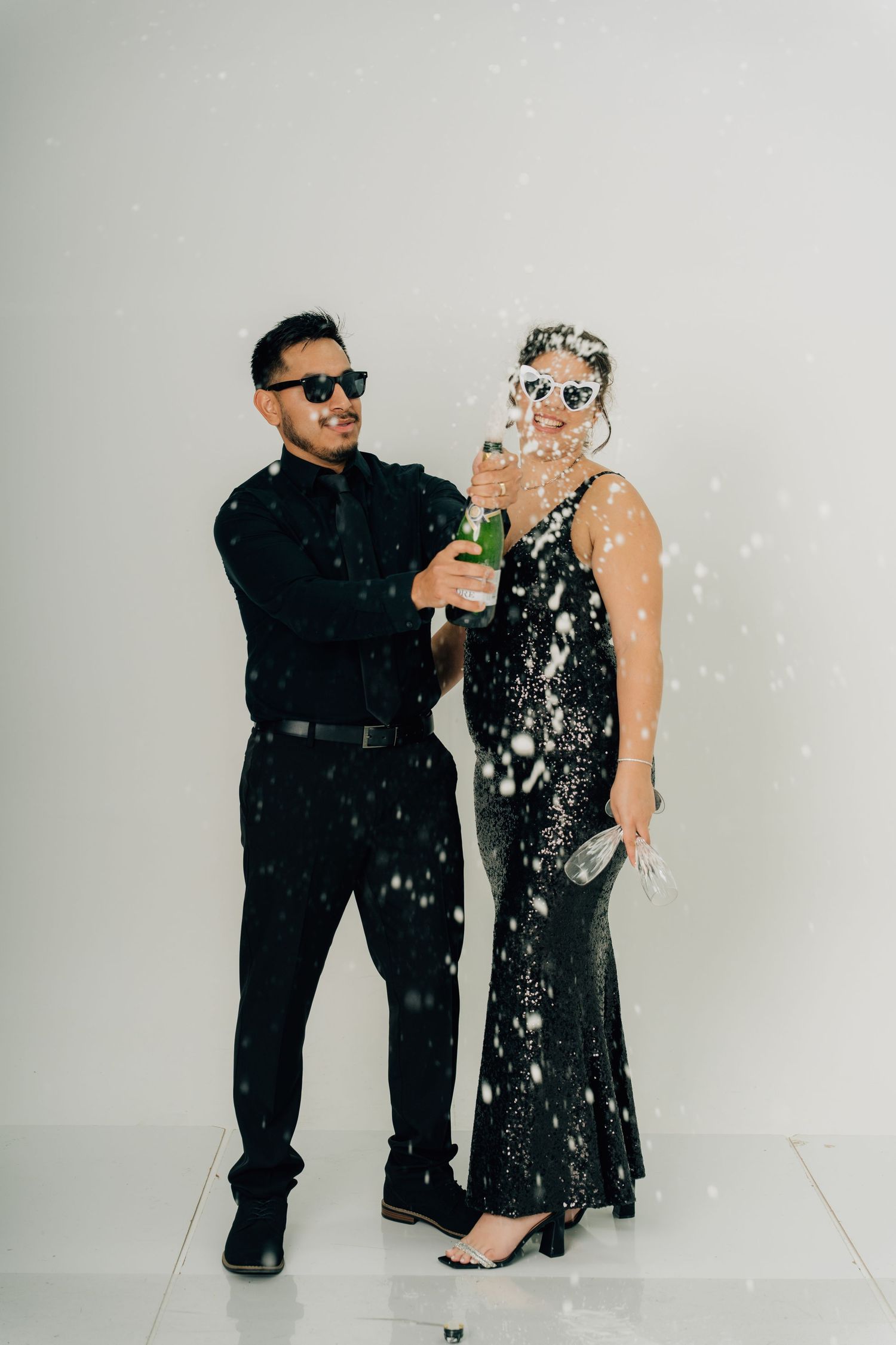 Stylish couple in formal attire posing with confetti against white backdrop.