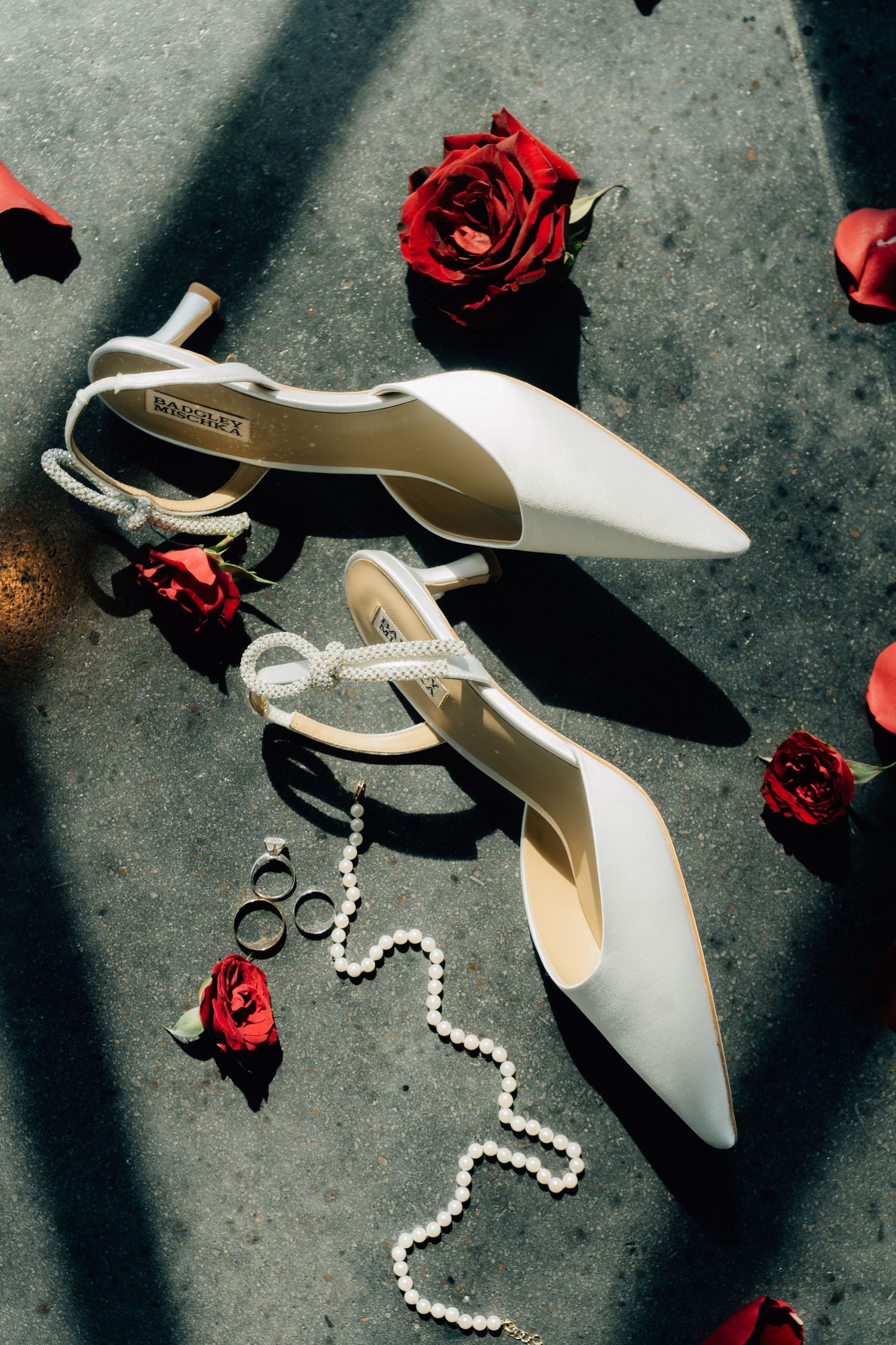 Wedding accessories including white heels, pearls and red rose petals artistically arranged.