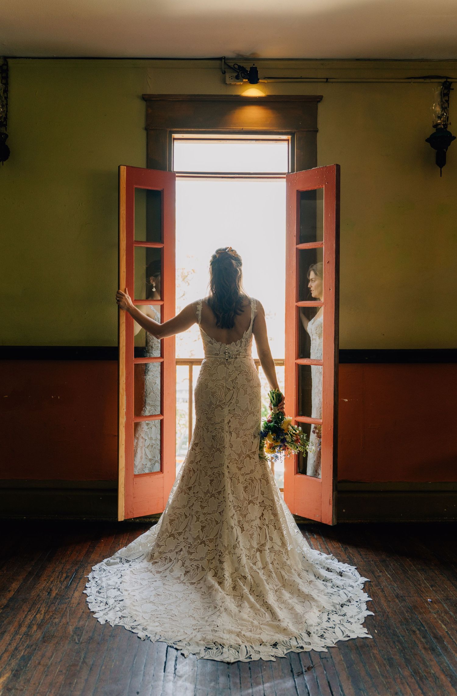 Elegant white wedding dress with flowing train silhouetted against bright windows in a vintage interior.