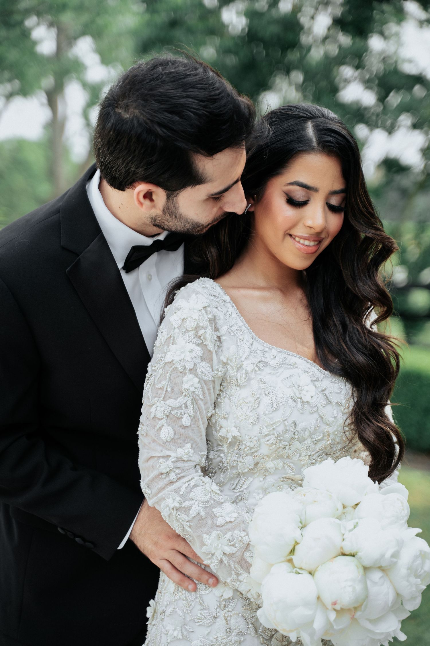 Elegant wedding couple embraces while bride holds white bouquet in lace long sleeve dress against green outdoor backdrop.
