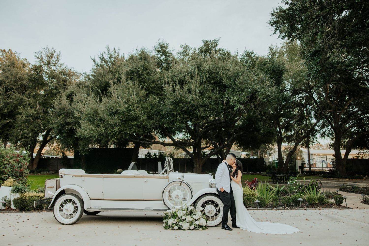 A romantic couple poses in an olive grove garden with a vintage silver car parked in the background.