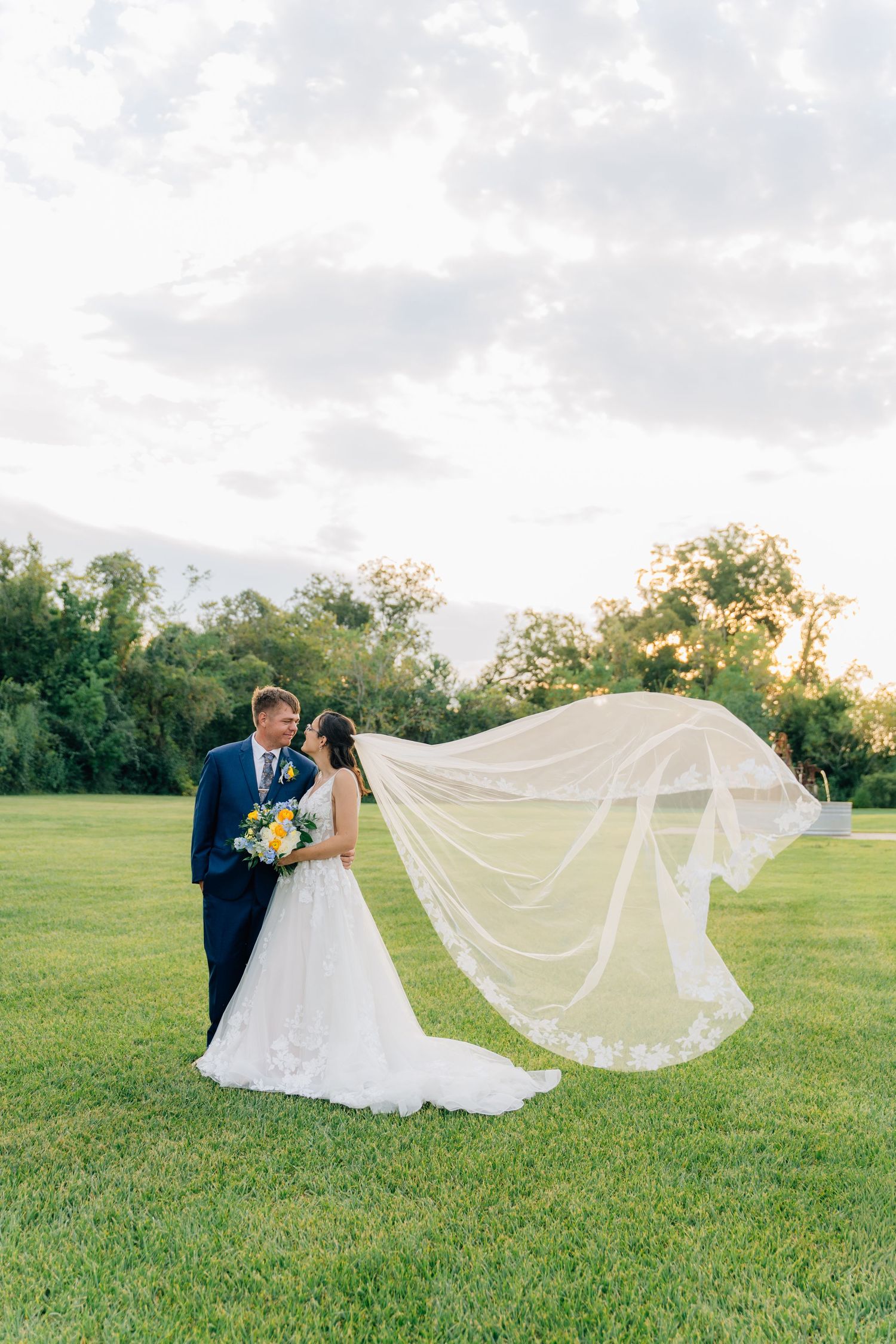 Scenic wedding photos in a green field with flowing veil creating dramatic movement at sunset.