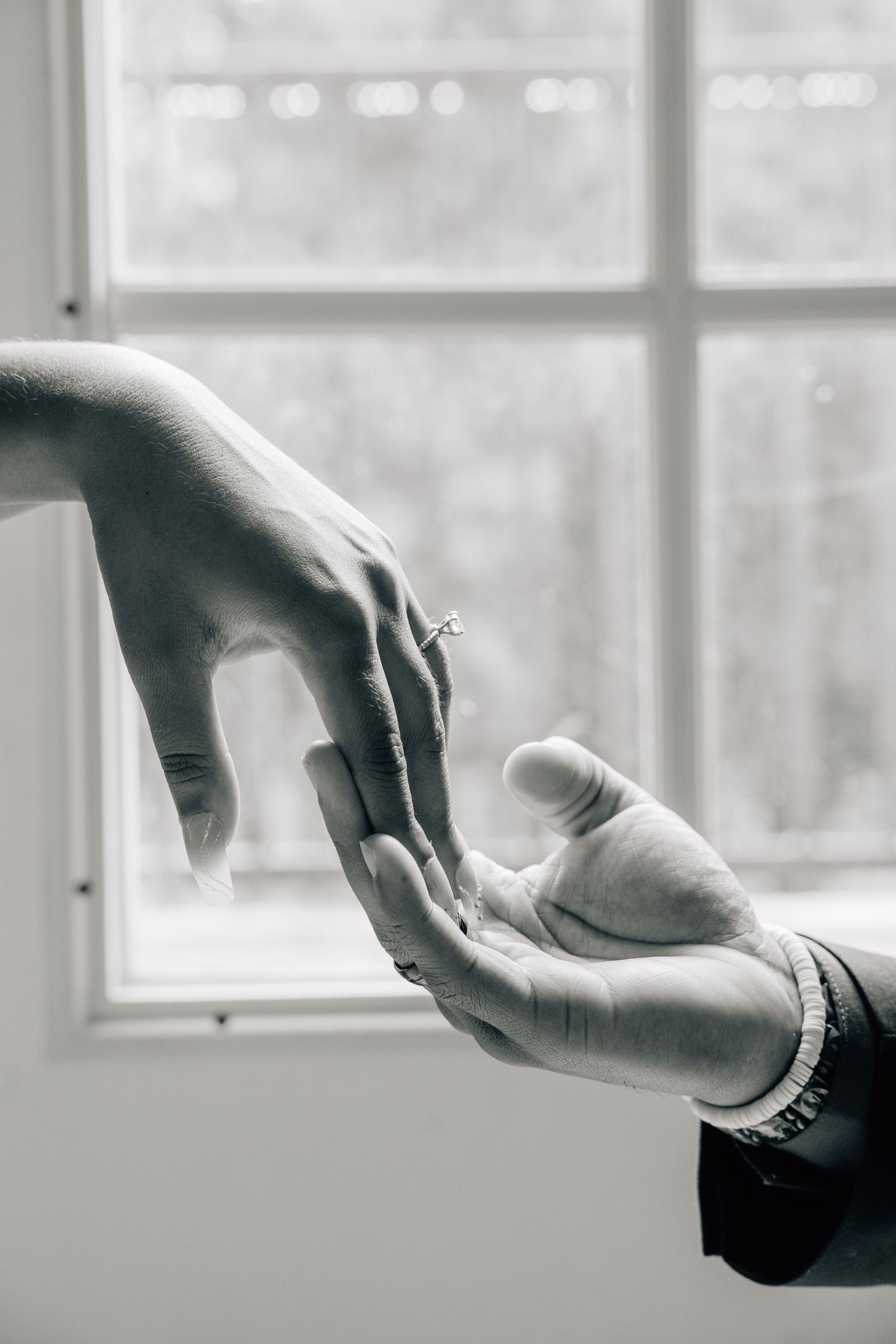 Two hands reaching towards each other in front of a bright window in black and white.