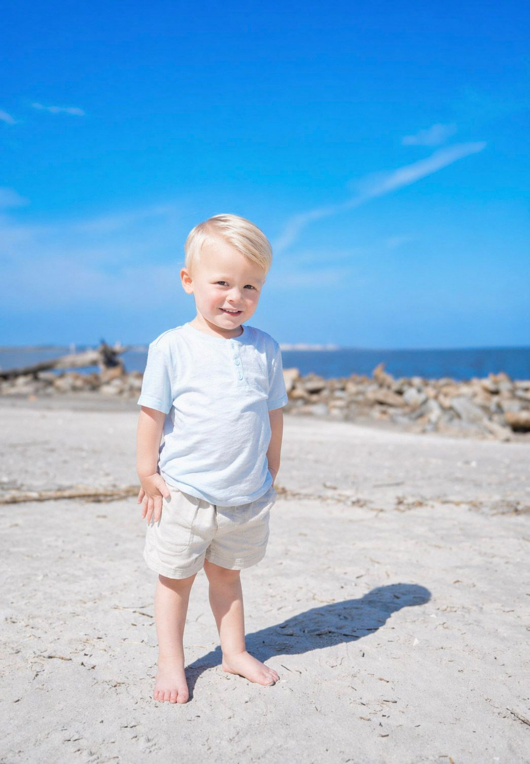 A smiling blonde toddler boy stands barefoot on a sandy beach wearing a light blue shirt and white shorts on a sunny day.