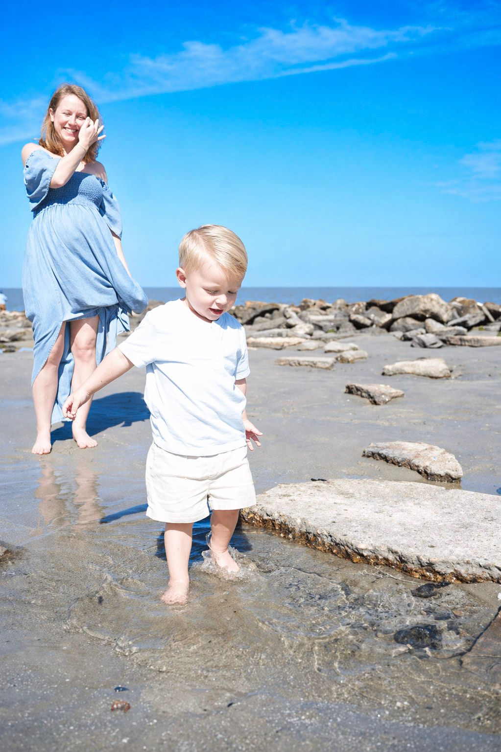 A toddler boy wades in shallow beach water while his mother watches nearby, with rocks and blue sky behind them.