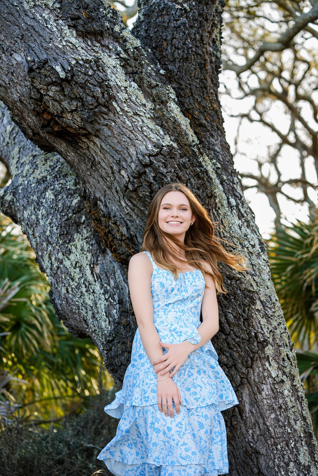 Young woman in blue floral dress smiling beside a large moss-covered tree outdoors.