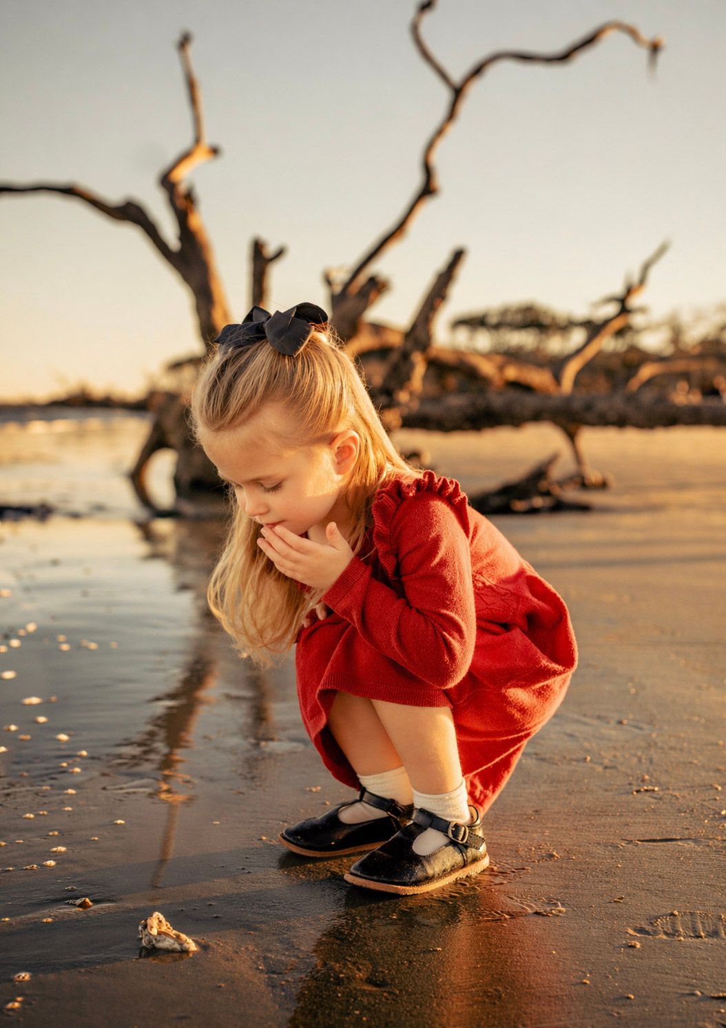 A young blonde girl in a red dress crouches on a wet beach at sunset, examining something in the sand.