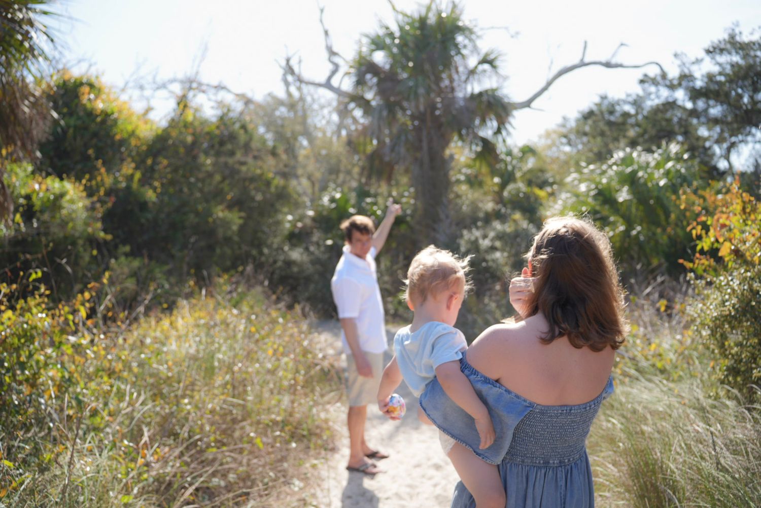 Family of three walking on a sunny sandy path surrounded by lush greenery and palm trees.