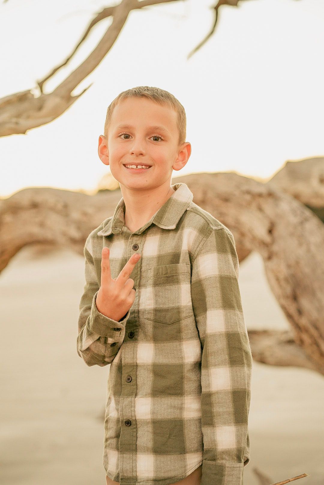 A smiling young boy in a plaid flannel shirt flashing a peace sign on a sandy beach with driftwood.