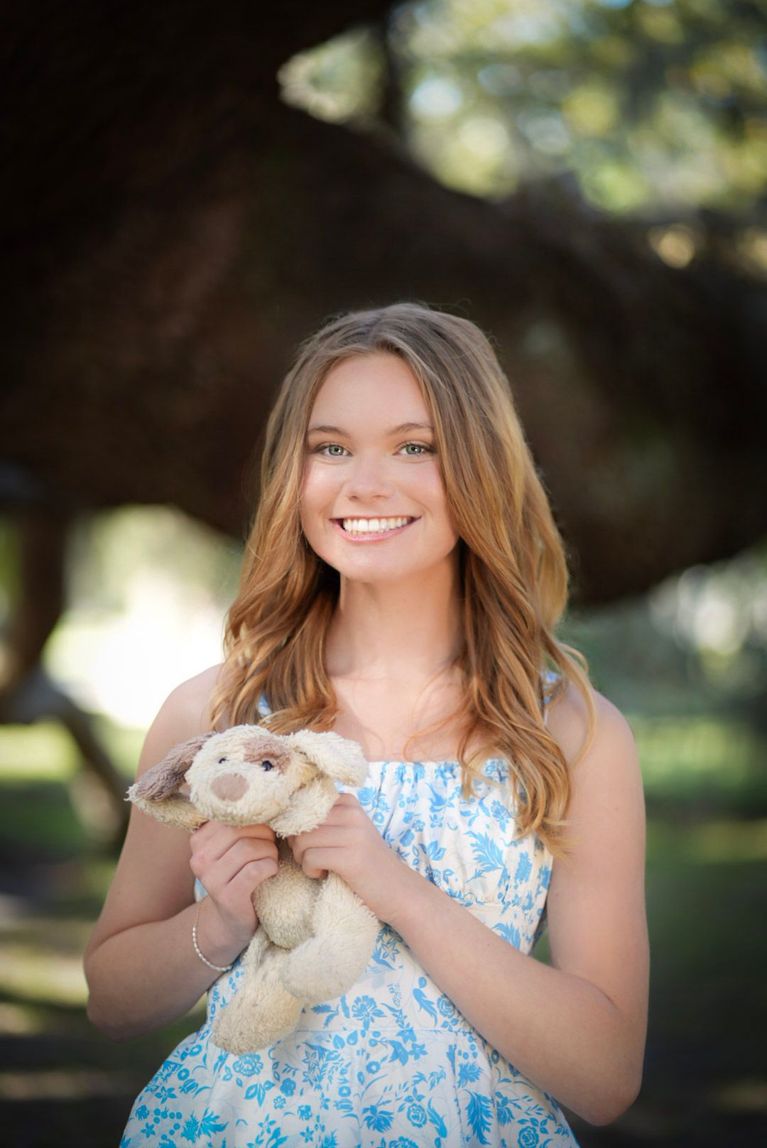 Smiling teenage girl holds stuffed bunny toy outdoors under large tree in blue dress.
