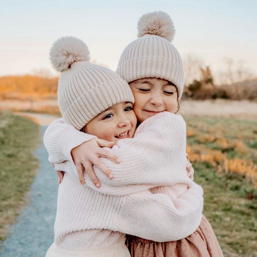 Two children in winter knit hats sharing a warm embrace against a soft natural backdrop.