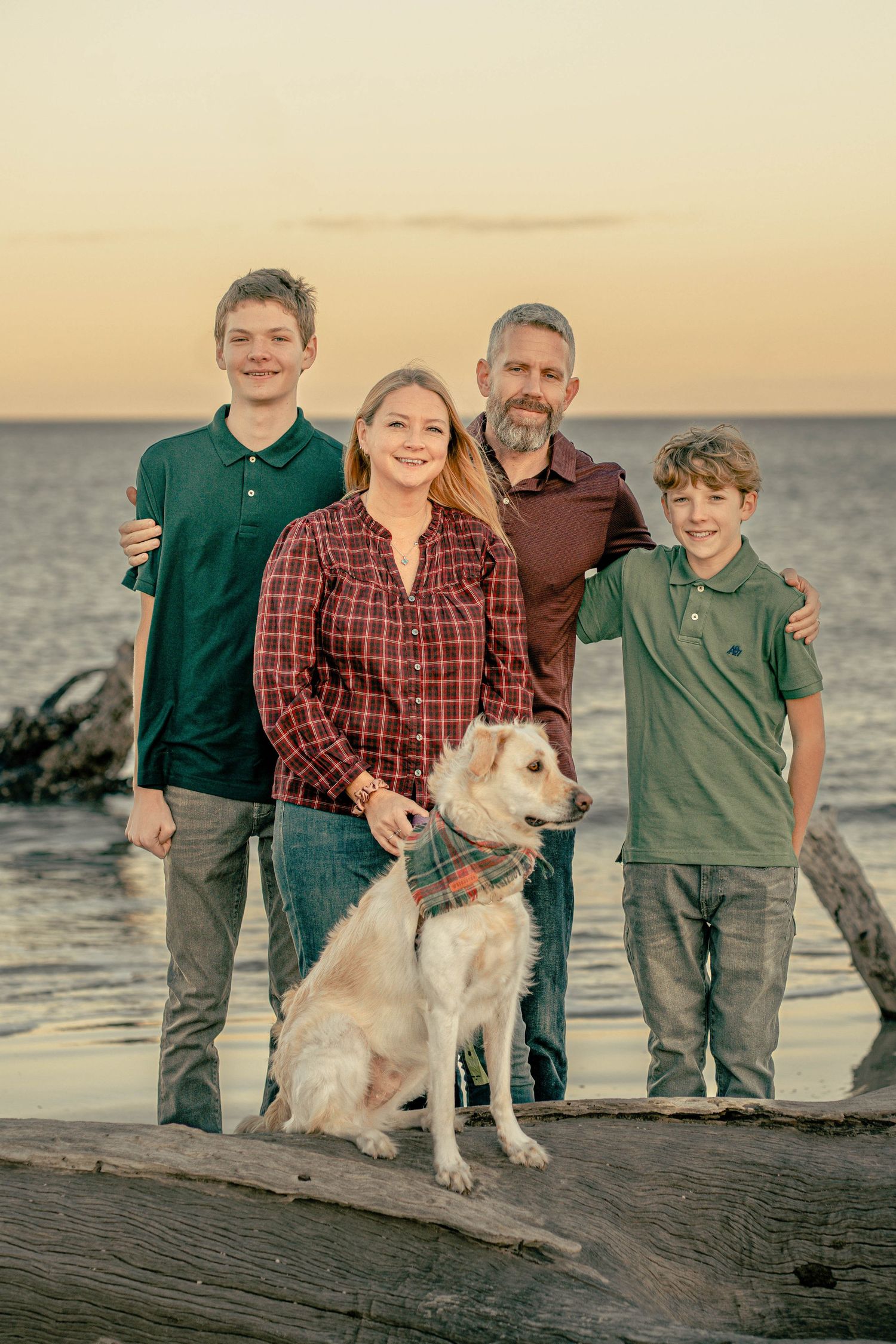 A family and their dog pose together on a beach at sunset near the water.