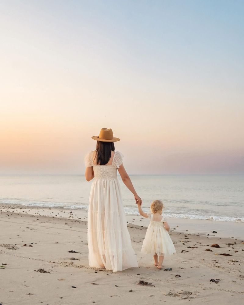 Mother and child in flowing white dresses walk together on peaceful beach.