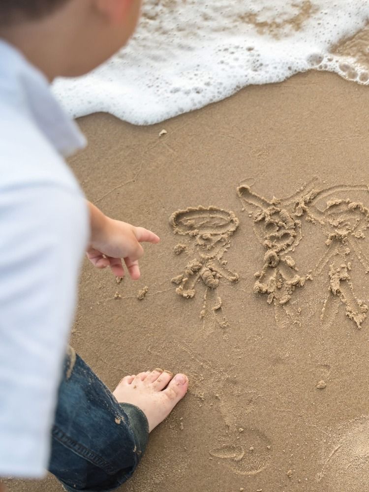 Child's hands drawing in wet sand at the beach with ocean waves in background.