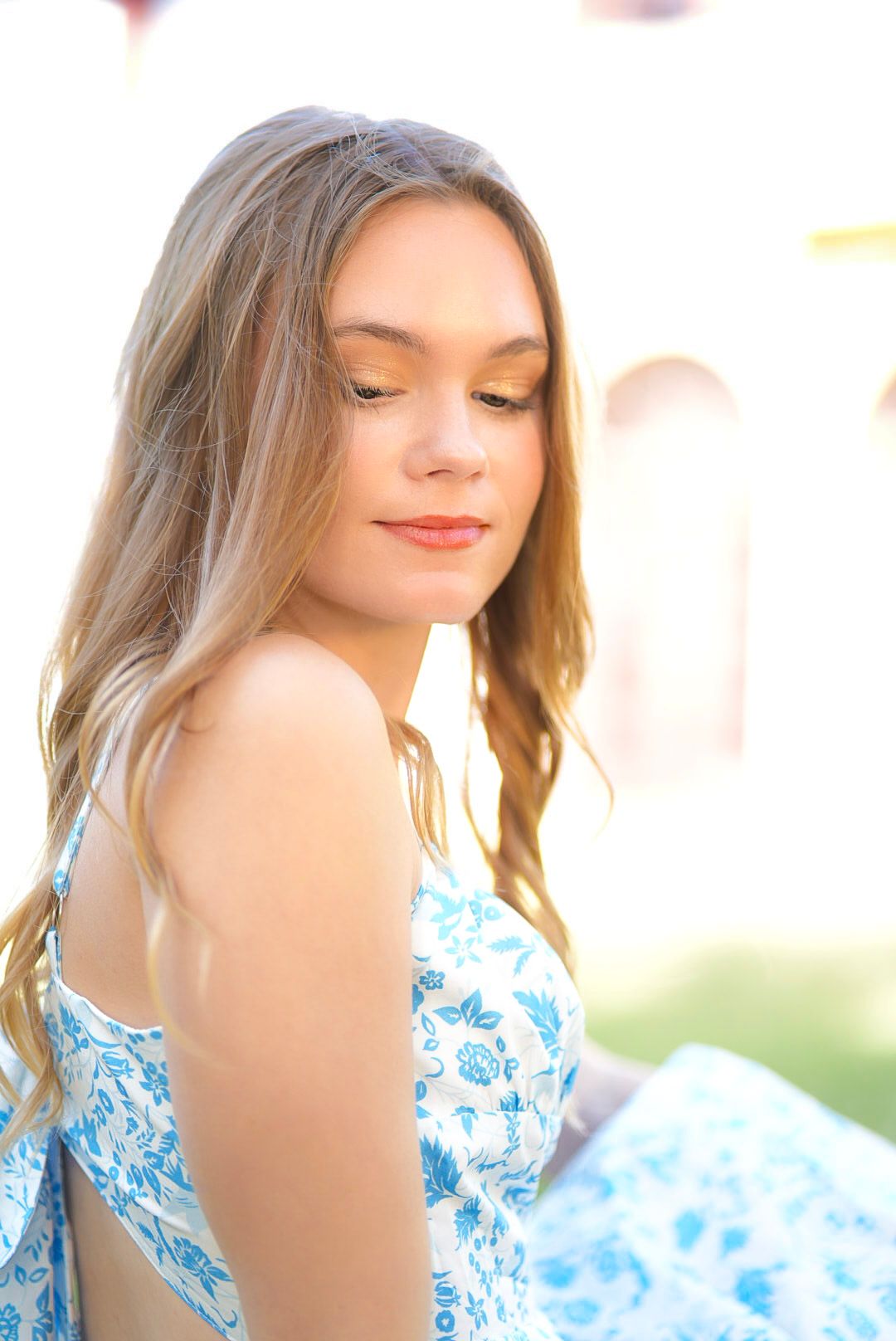 Young woman with long blonde hair wearing a blue floral sundress, looking down in soft natural sunlight outdoors.