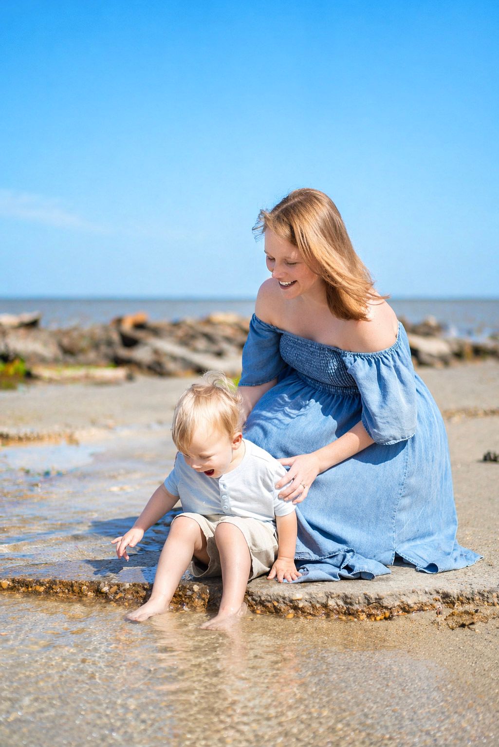 A smiling mother in a blue dress sits with her toddler on a sunny beach, watching the child play in shallow water.