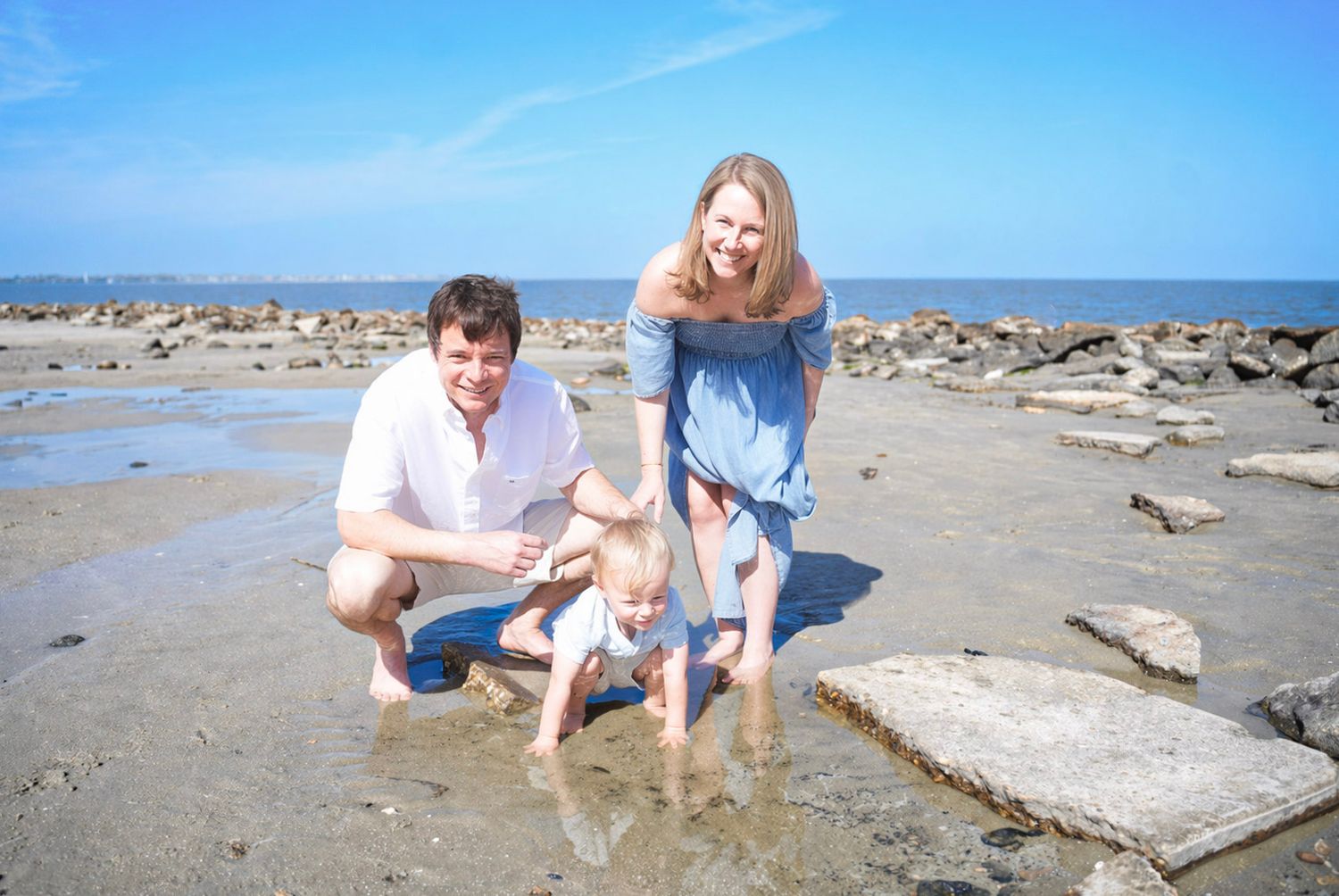 Family of three enjoying a sunny beach day, parents crouching beside toddler near tidal pools on rocky shore.