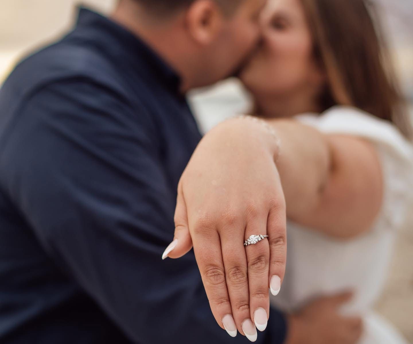 Close-up of engagement ring on hand with couple embracing in background.