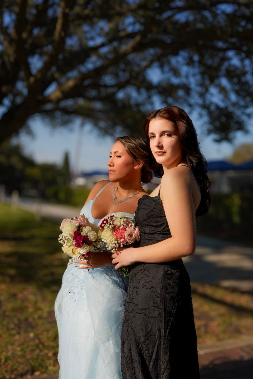 Two elegant women in formal gowns pose outdoors, one holding a bouquet of pink and white flowers under a shaded tree.