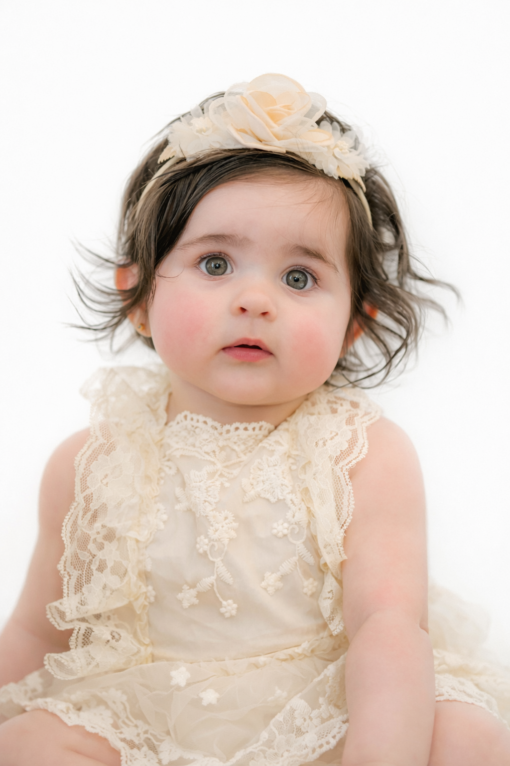 Adorable baby girl in cream lace dress and floral headband with dark hair and big eyes on white background.
