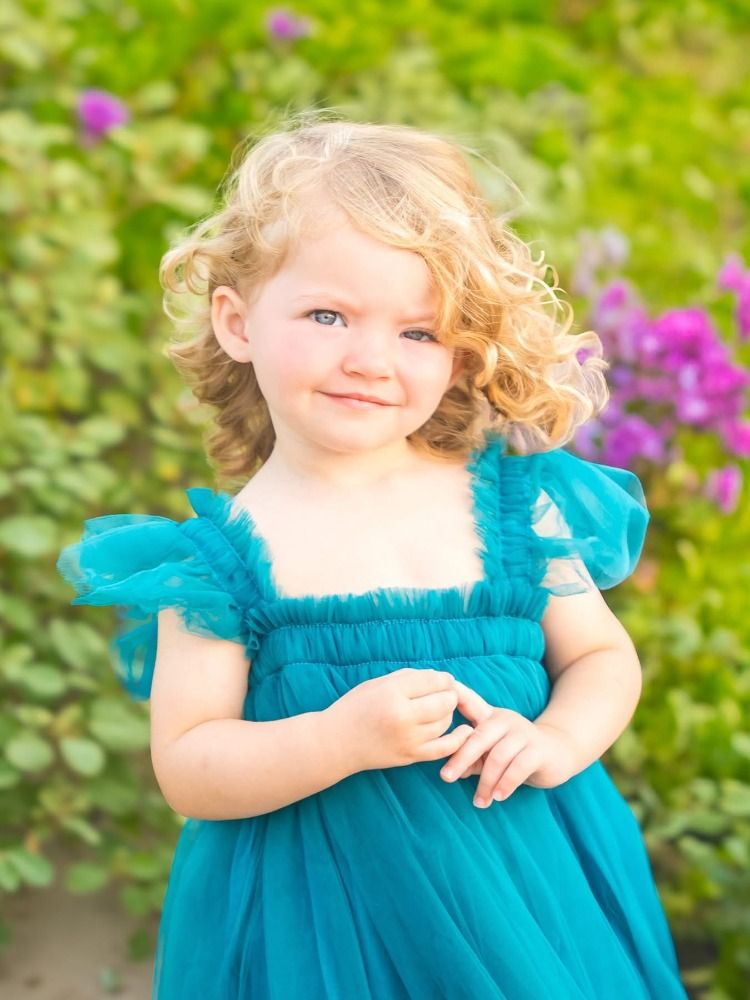 Child in ruffled turquoise dress poses in garden with blooming pink flowers.