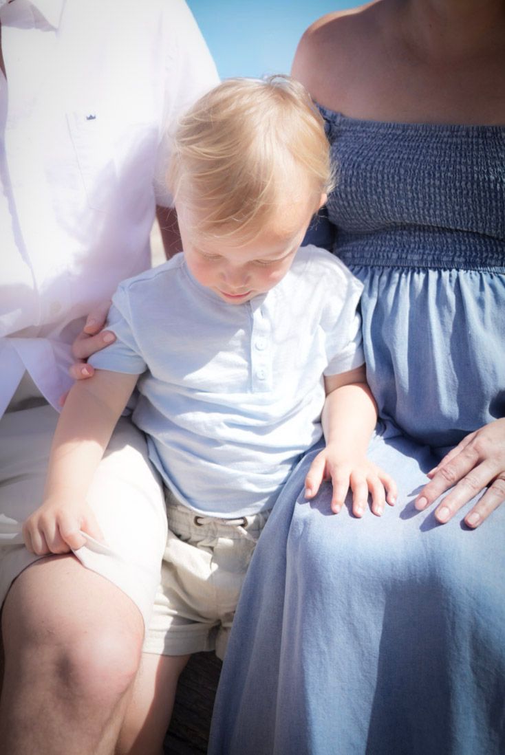 Toddler boy in light blue shirt sitting between parents outdoors on a sunny day.