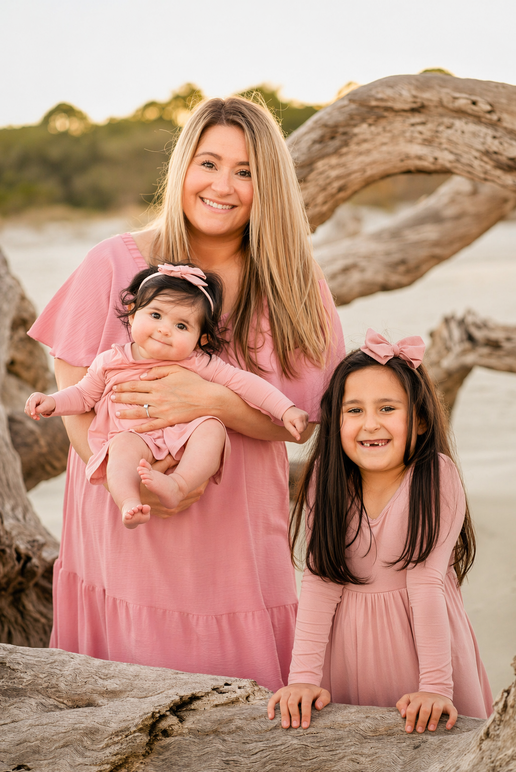 A smiling woman in a pink dress holds a baby girl while an older girl in pink poses beside her on a beach.