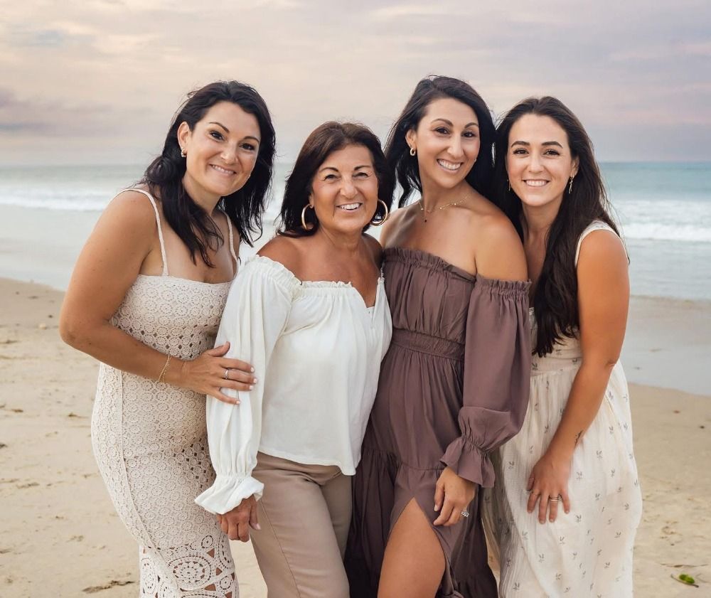 Group of women in neutral dresses smile together on a beach at sunset.