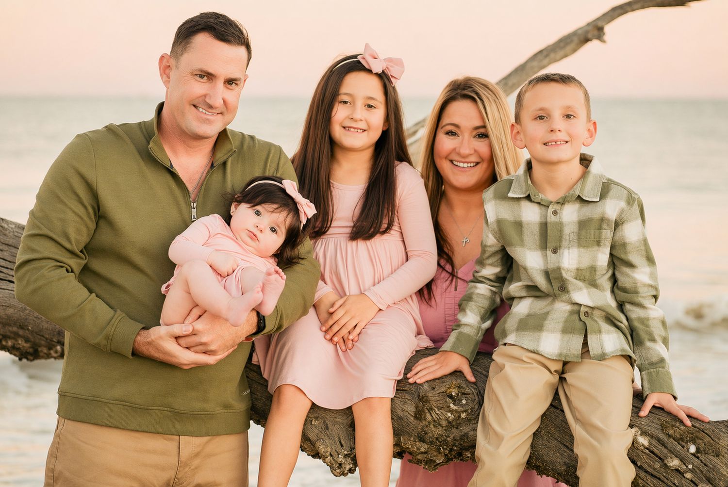 Family of five smiling together on a beach at sunset, parents with three children in coordinating outfits.